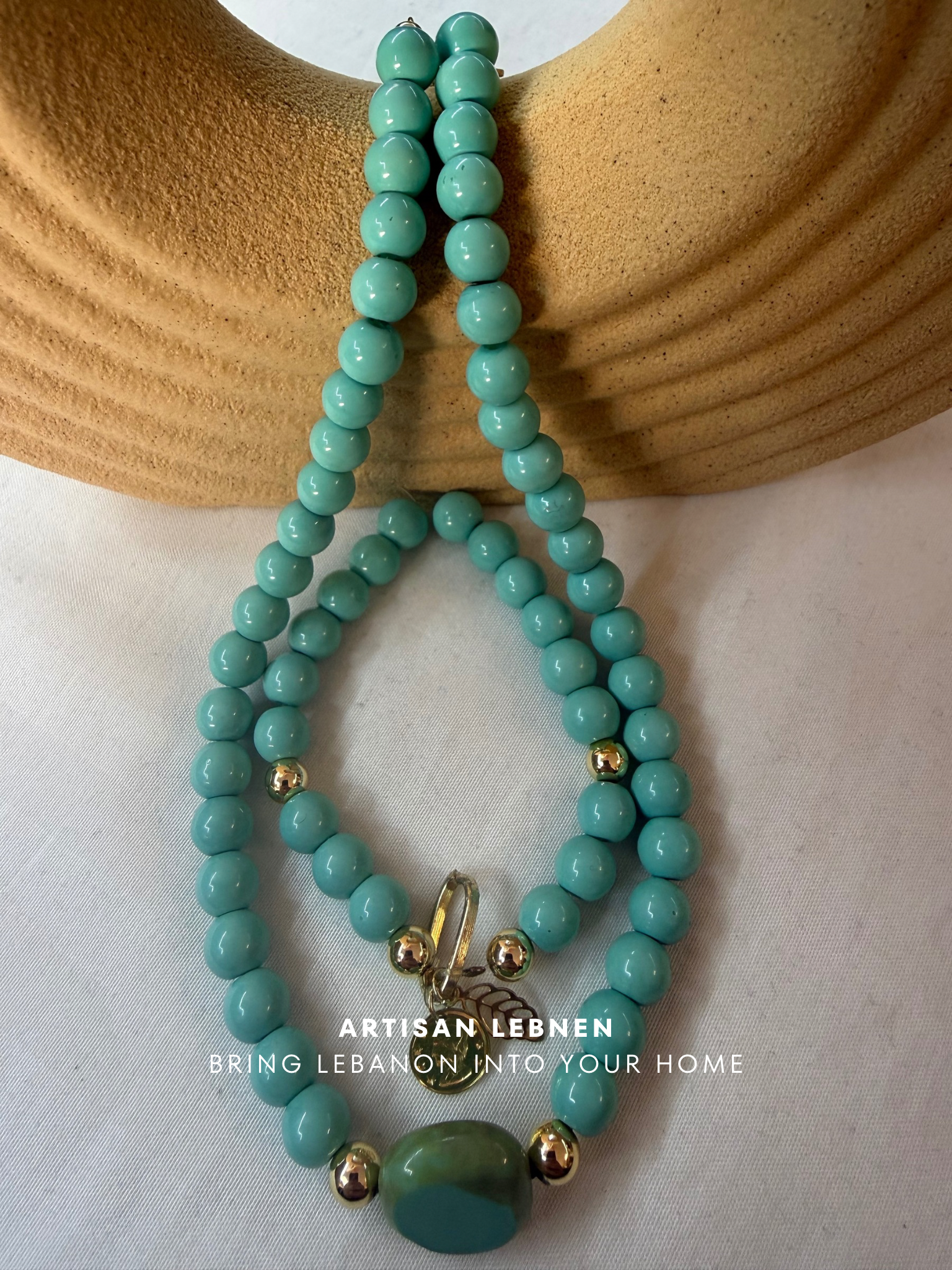 Turquoise beaded necklace with gold accents on a beige surface, featuring 'Artisan Lebanon' branding.