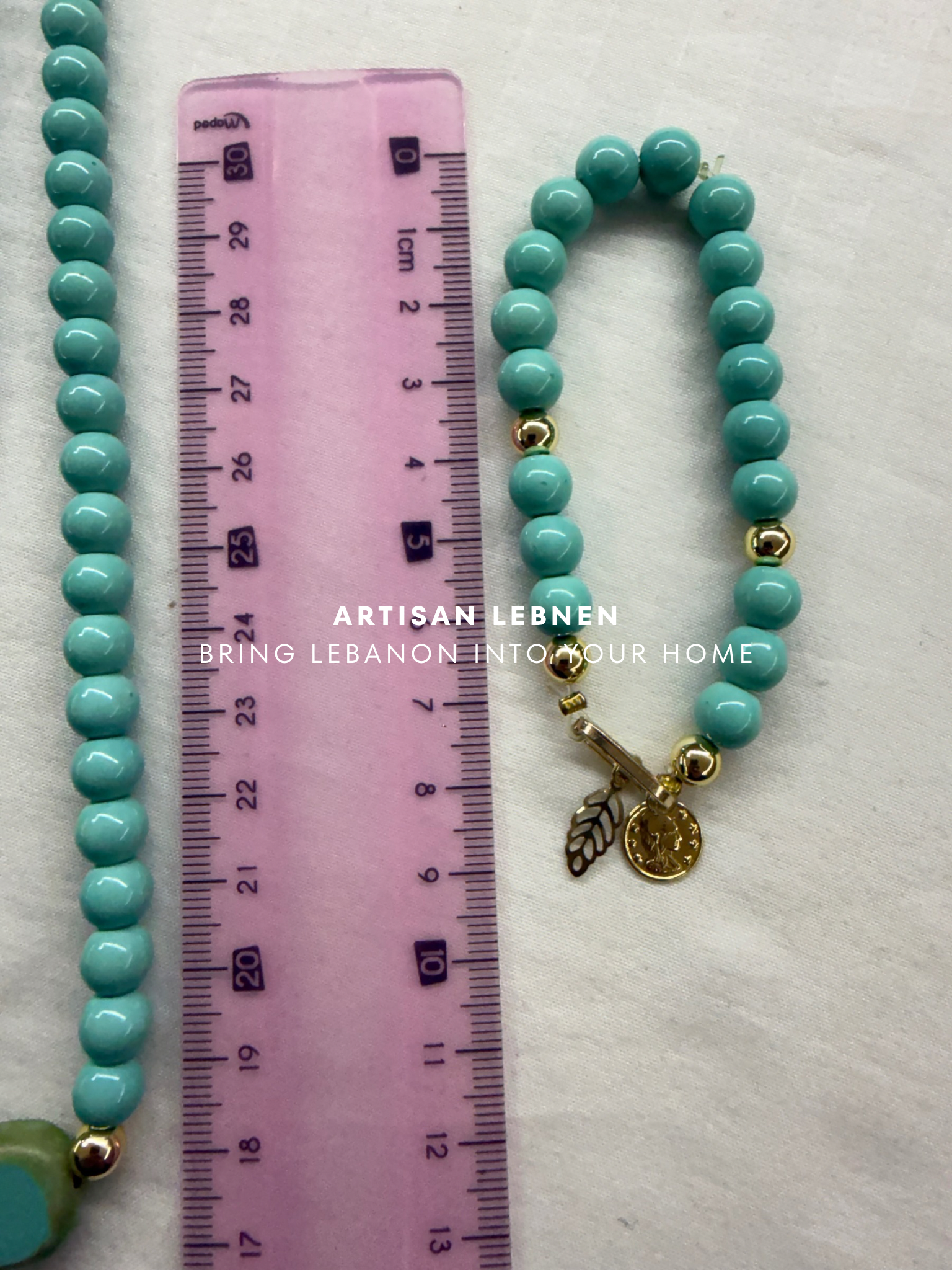 Turquoise beaded bracelet with a gold charm next to a pink ruler on a white background.