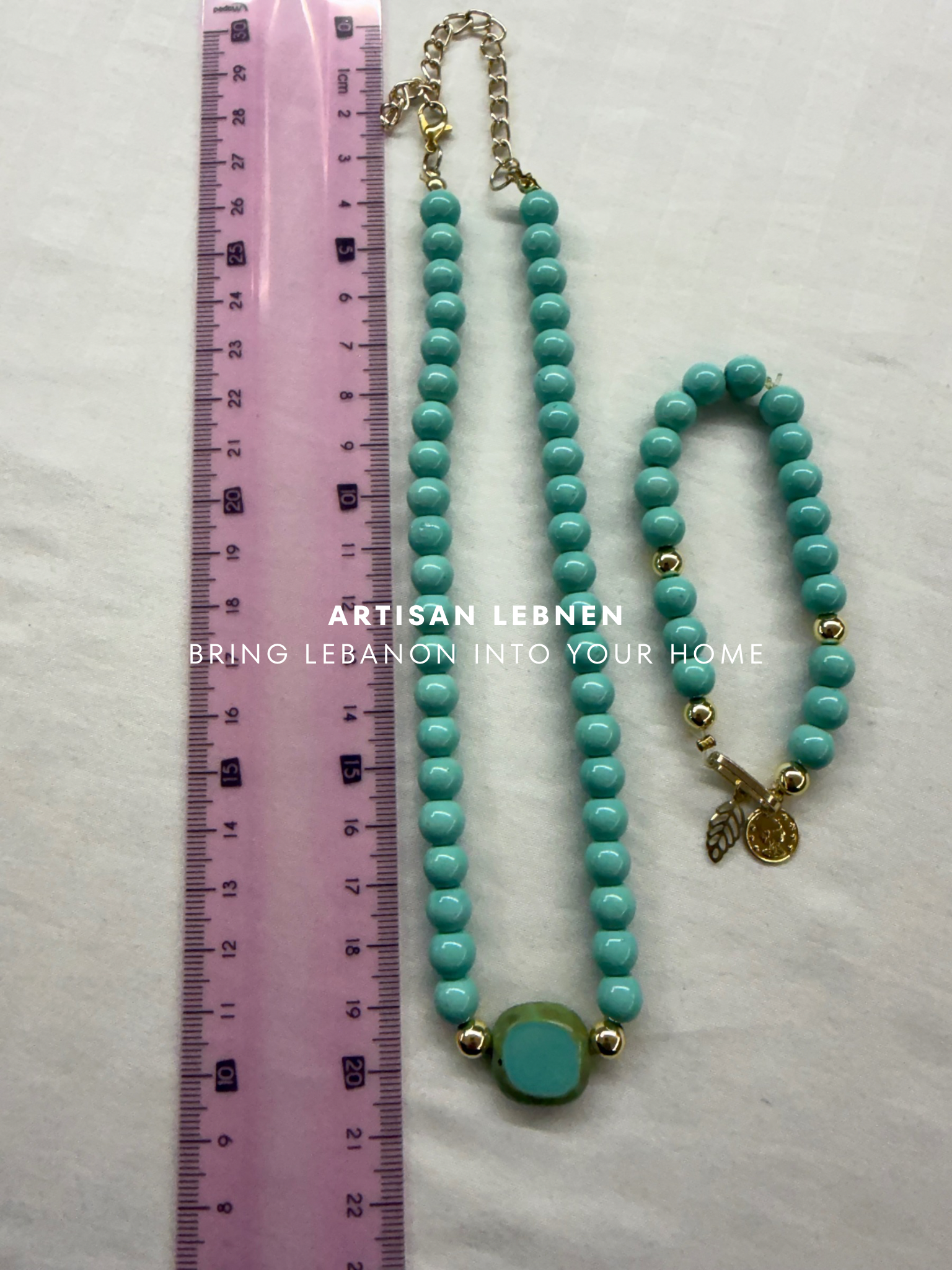 Turquoise beaded necklace with gold accents next to a pink ruler on a white background, with 'Artisan Lebnen' branding.