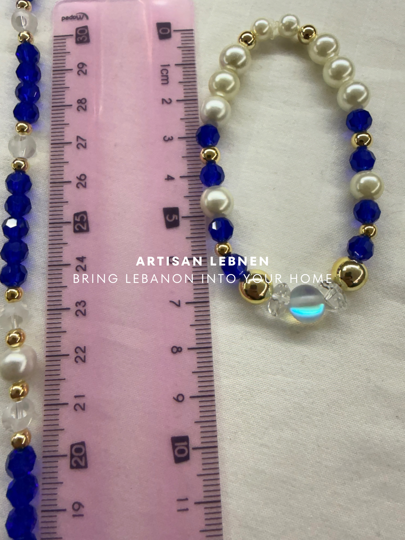 Beaded bracelet with blue, white, and gold beads on a ruler for size reference, with 'Artisan Lebnen' branding.